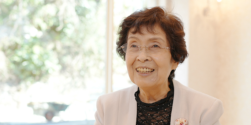 高齢女性が施設内で笑顔でくつろぐ様子。窓から自然光が入る明るい空間。